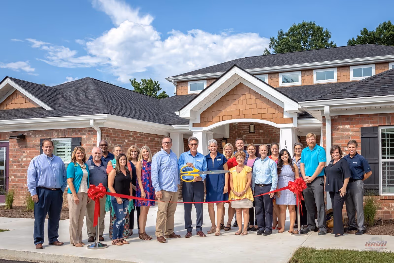 A group of people standing outside a brick building with a black roof, participating in a ribbon-cutting ceremony. The group is holding a red ribbon stretched across the entrance, and a man in the center is holding large ceremonial scissors. The sky is blue with some clouds, and there are trees in the background.