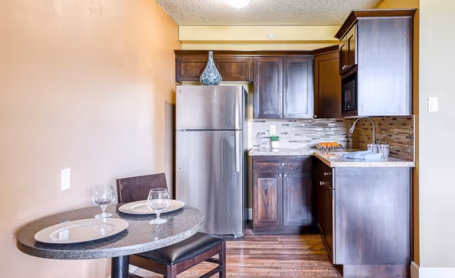 Small kitchen area with dark wood cabinets, a stainless steel refrigerator, a countertop with a sink, and a small round dining table set with two plates and two wine glasses, accompanied by two chairs.