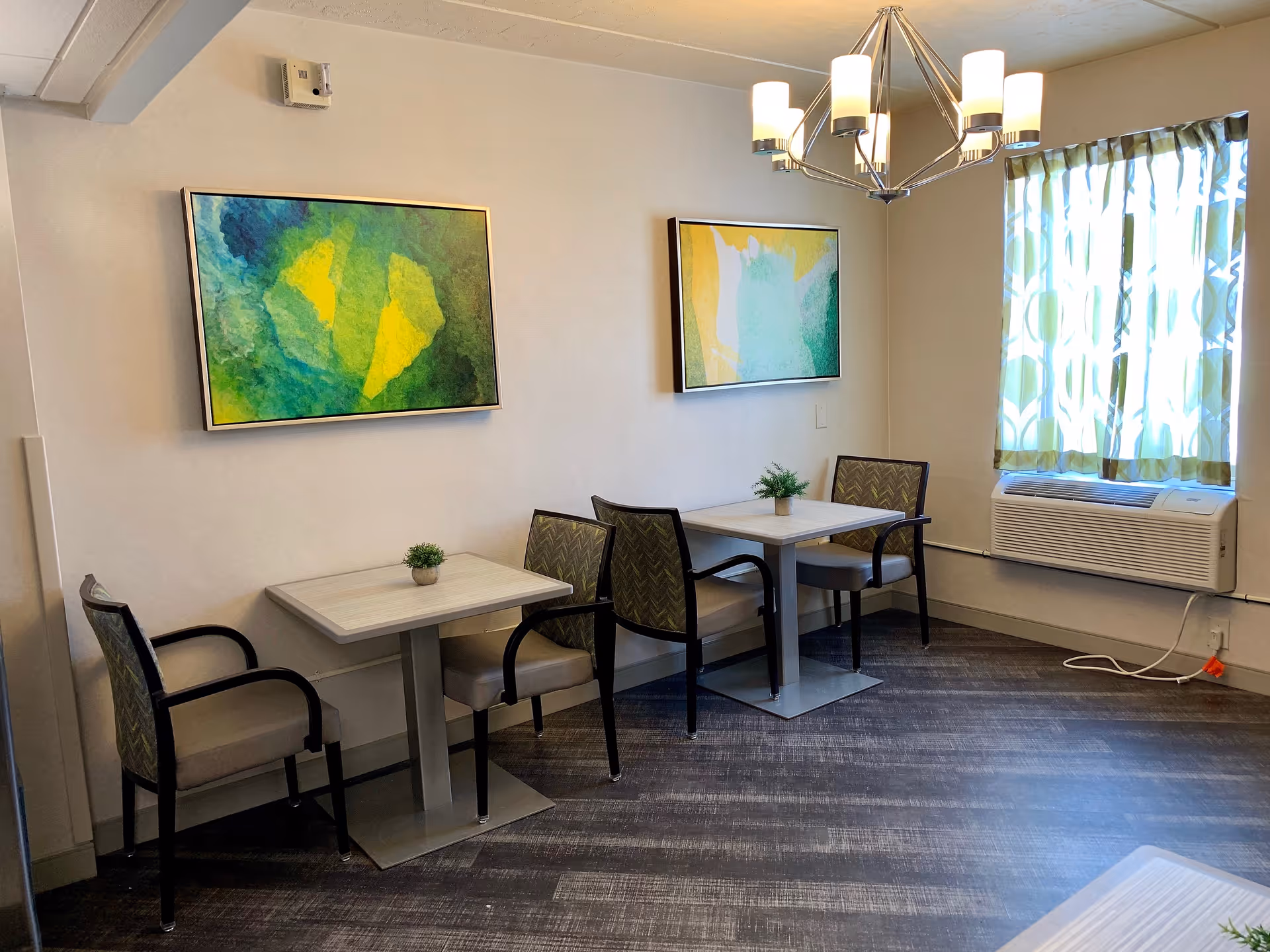 A small dining area with two square tables, each accompanied by two chairs with patterned upholstery. Two abstract paintings with green and yellow tones hang on the wall above the tables. A window with patterned curtains and an air conditioning unit below it is on the right side. A modern chandelier with six lights hangs from the ceiling.