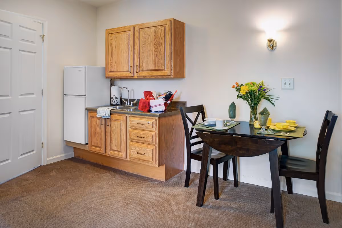 A small kitchenette area with wooden cabinets, a countertop with a sink, and a white refrigerator. Next to the kitchenette is a dark wooden dining table set for two with plates, cups, and a vase of colorful flowers. The room has beige walls and carpeted flooring.