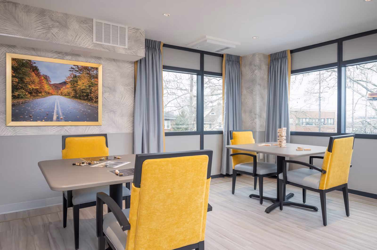 A bright and modern room with two tables and four yellow cushioned chairs. The tables have board games on them, including a Jenga tower. Large windows with gray curtains allow natural light to fill the space. A framed picture of a road lined with autumn trees hangs on the wall.