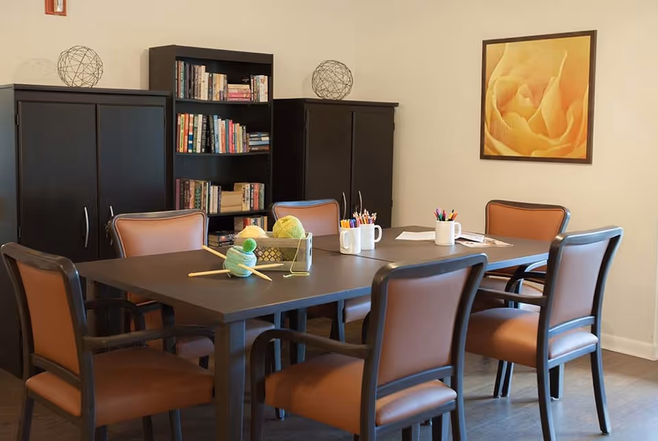A communal activity room with a rectangular table surrounded by six chairs, craft supplies and yarn on the table, and bookshelves and cabinets along the wall.