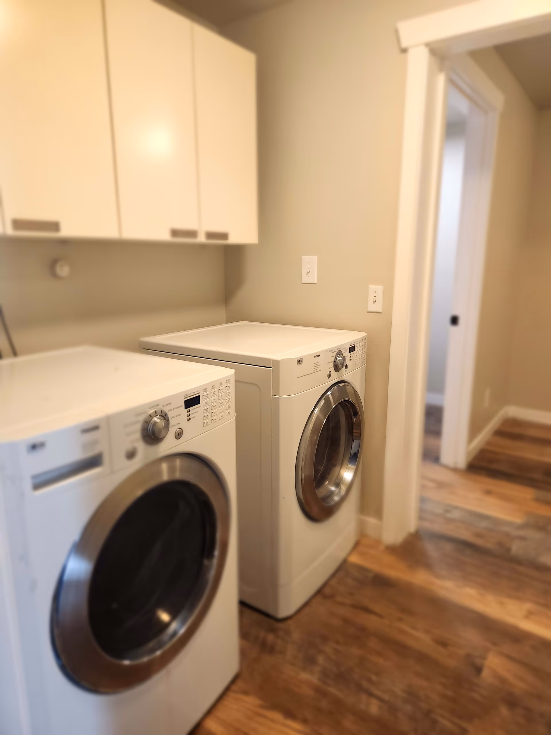 Laundry room with a white front-loading washing machine and dryer side by side, white cabinets mounted on the wall above them, and wooden flooring. A doorway leads to another room with similar flooring.