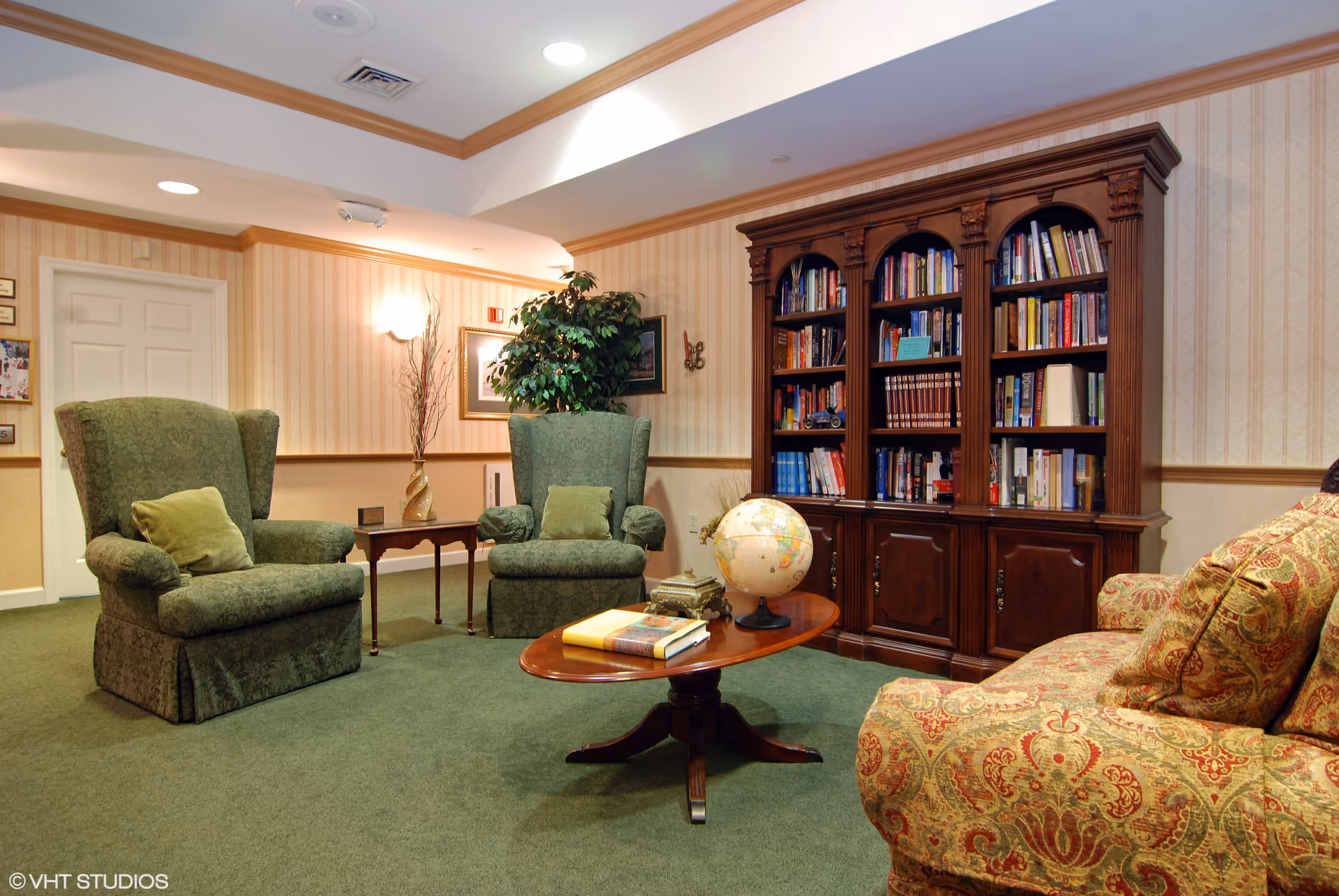 A cozy living room area with two green upholstered armchairs, a patterned sofa, a wooden coffee table with books and a globe, and a large wooden bookshelf filled with books. The room has green carpet, wallpapered walls with a chair rail, and soft lighting.