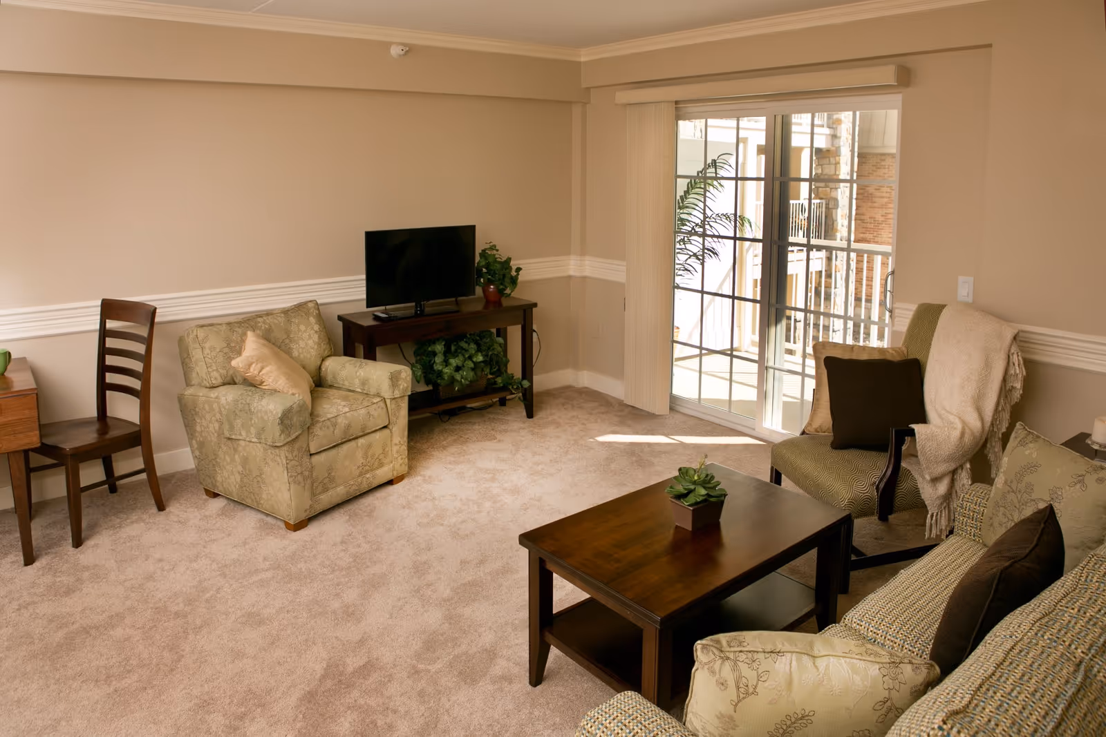 A cozy living room with beige walls and carpet, featuring a patterned armchair with a pillow, a wooden chair, a small TV on a wooden stand with plants underneath, a wooden coffee table with a small potted plant, and a sofa with multiple cushions and a throw blanket. Sliding glass doors lead to an outdoor balcony with a view of a building exterior.