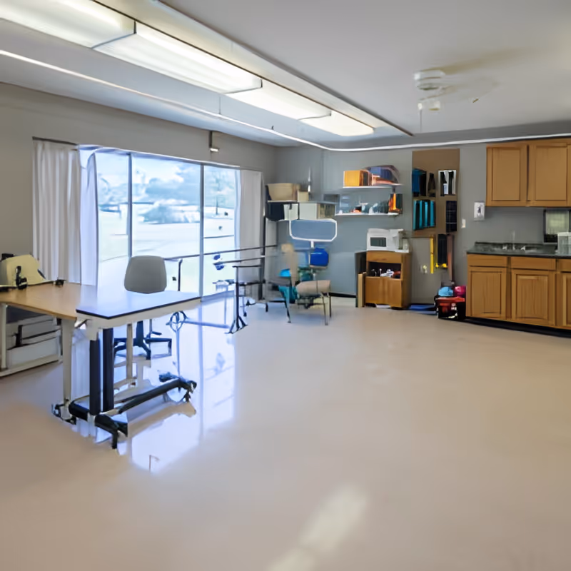 A spacious room with large windows covered by white curtains, a desk with a chair, medical or rehabilitation equipment, shelves with various items, and wooden cabinets with a countertop and sink.