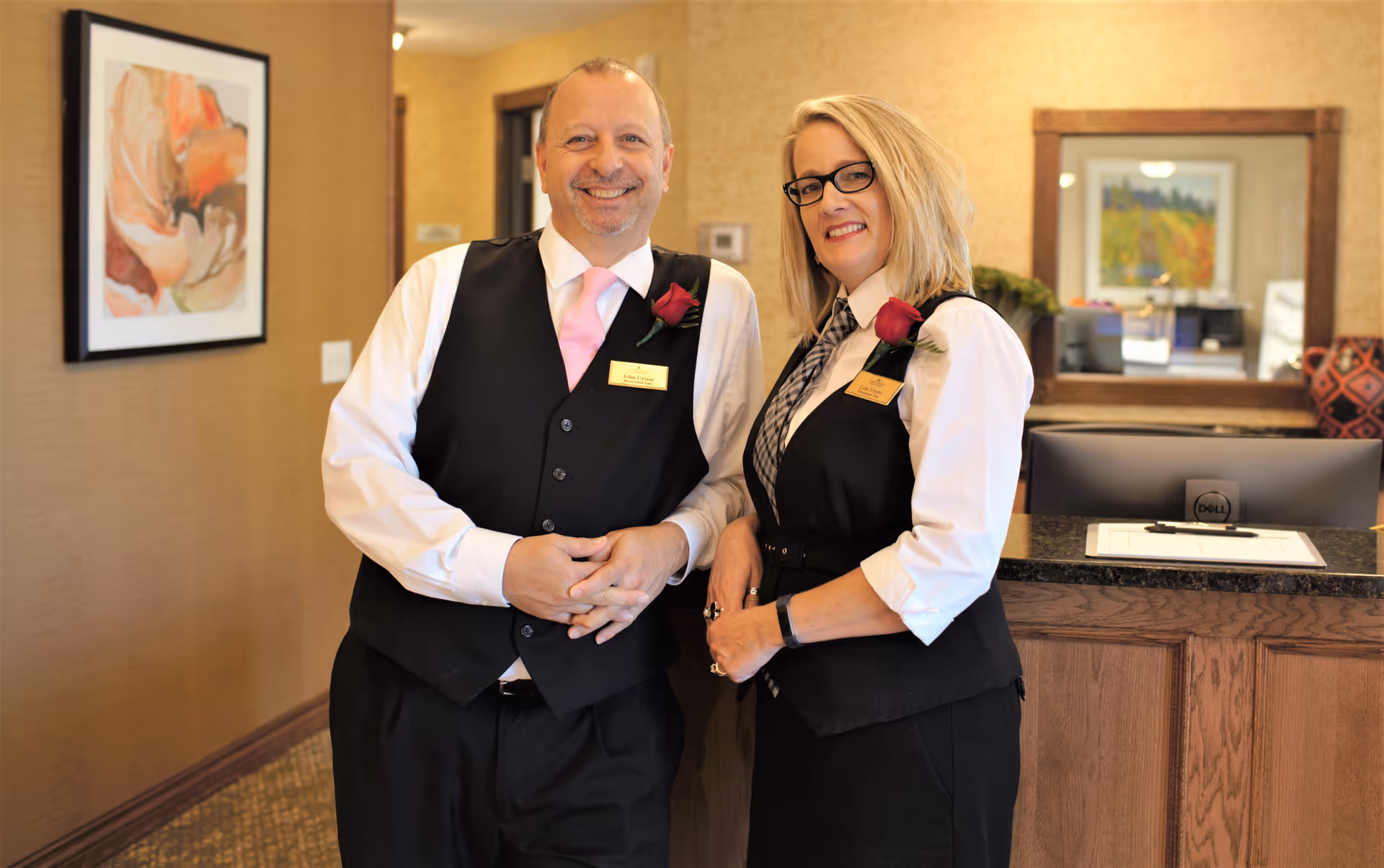 Two smiling staff members wearing black vests, white shirts, and name tags stand behind a wooden reception desk in a warmly decorated room with framed artwork on the walls.