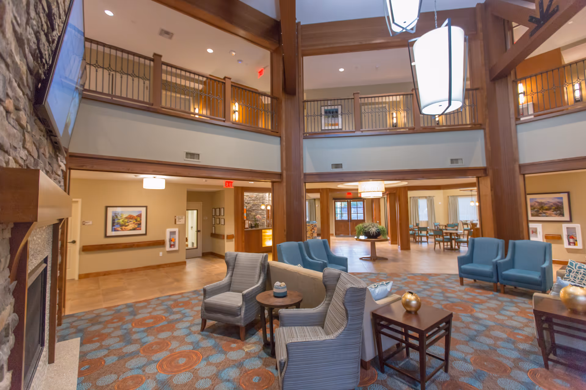 Spacious two-story senior living lobby with seating areas, exposed wooden beams, a stone fireplace, and a central chandelier.