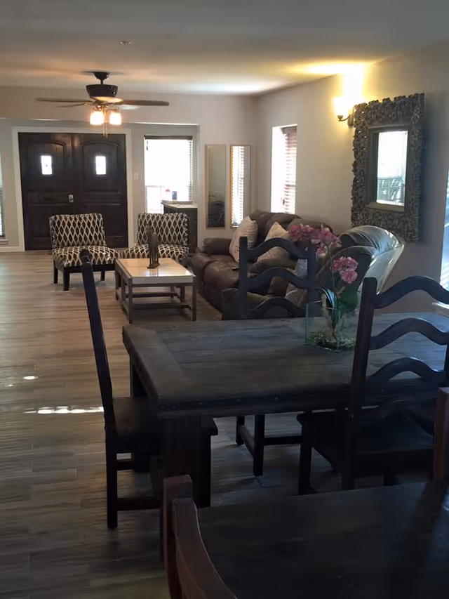 Interior view of a living room and dining area with wooden flooring. The living room features a brown leather sofa, two patterned armchairs, a wooden coffee table with a decorative item, and a large ornate mirror on the wall. The dining area has a dark wooden table with matching chairs and a vase with pink flowers. Ceiling fan with lights is visible, and there are windows with blinds letting in natural light.