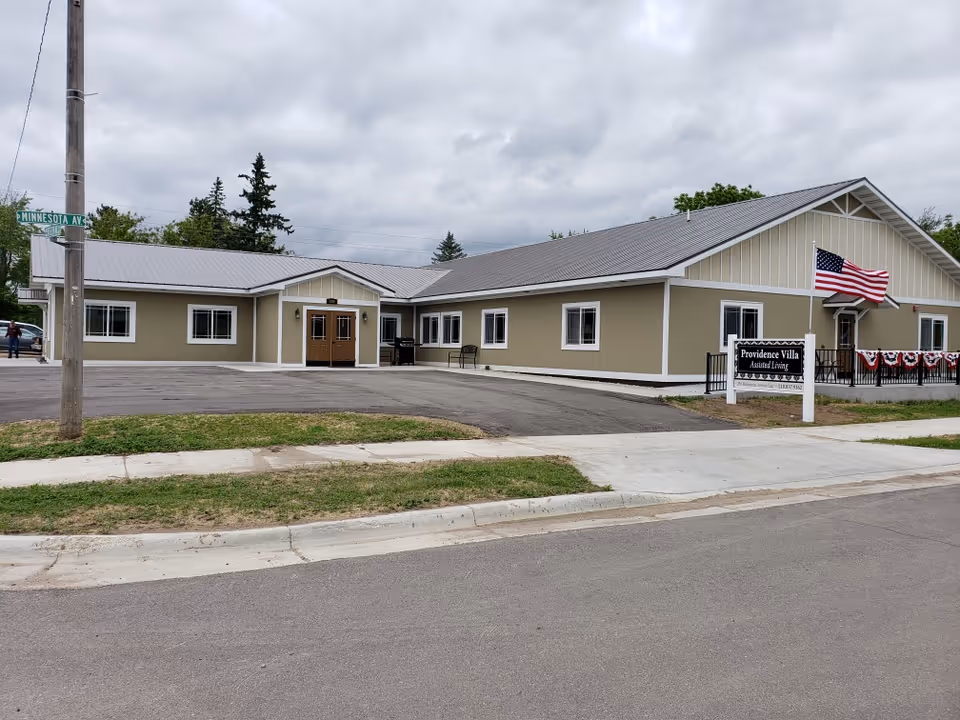 Exterior view of Providence Villa Assisted Living facility, a single-story building with beige walls and a gray metal roof. There is an American flag on a pole near the entrance, a sign with the facility name, and a street sign indicating Minnesota Ave. The sky is cloudy.