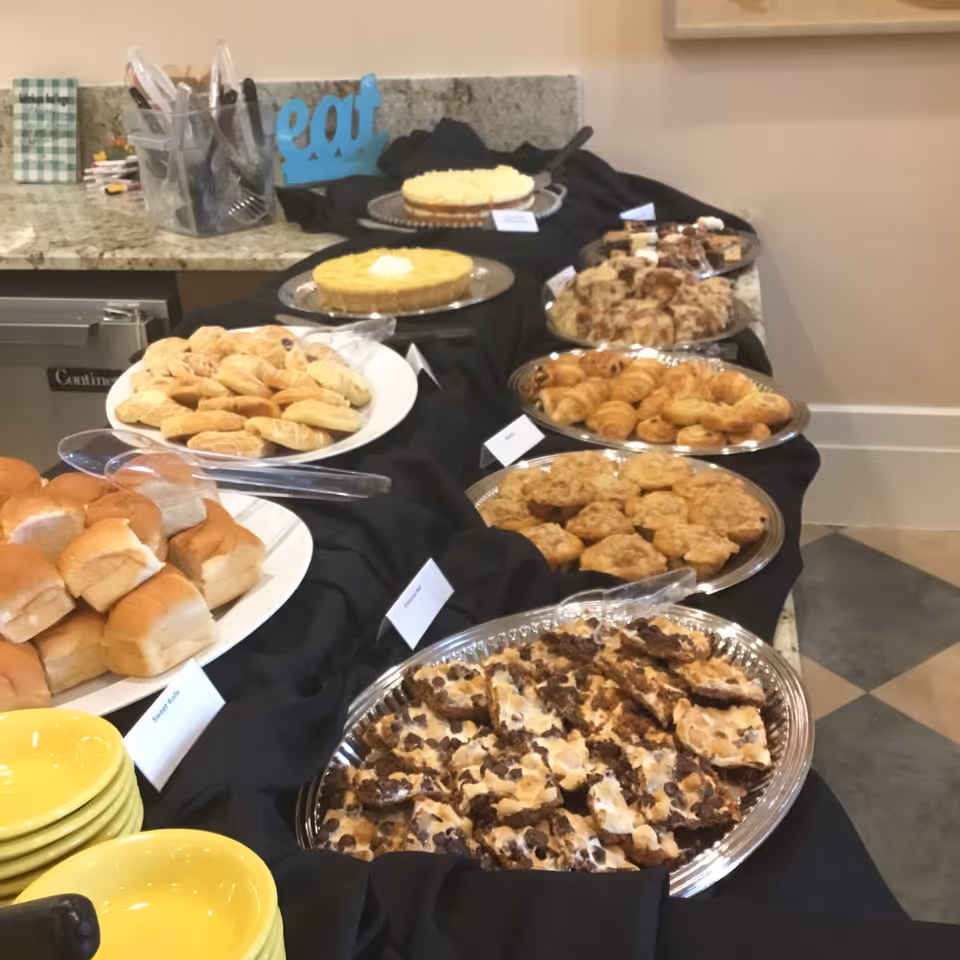A buffet table covered with a black cloth displaying various baked goods including rolls, cookies, croissants, and two types of pies. Yellow bowls are stacked at the front left corner of the table. Utensils and a blue decorative sign that says 'eat' are visible on the countertop behind the table.