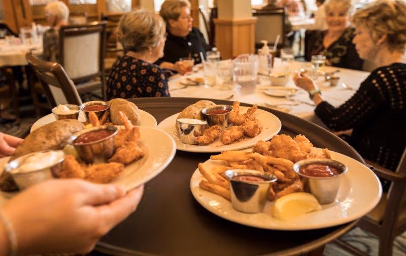 A server holding a tray with three plates of food including fried shrimp, French fries, baked potatoes, and dipping sauces in a dining room where elderly people are seated and conversing at tables.