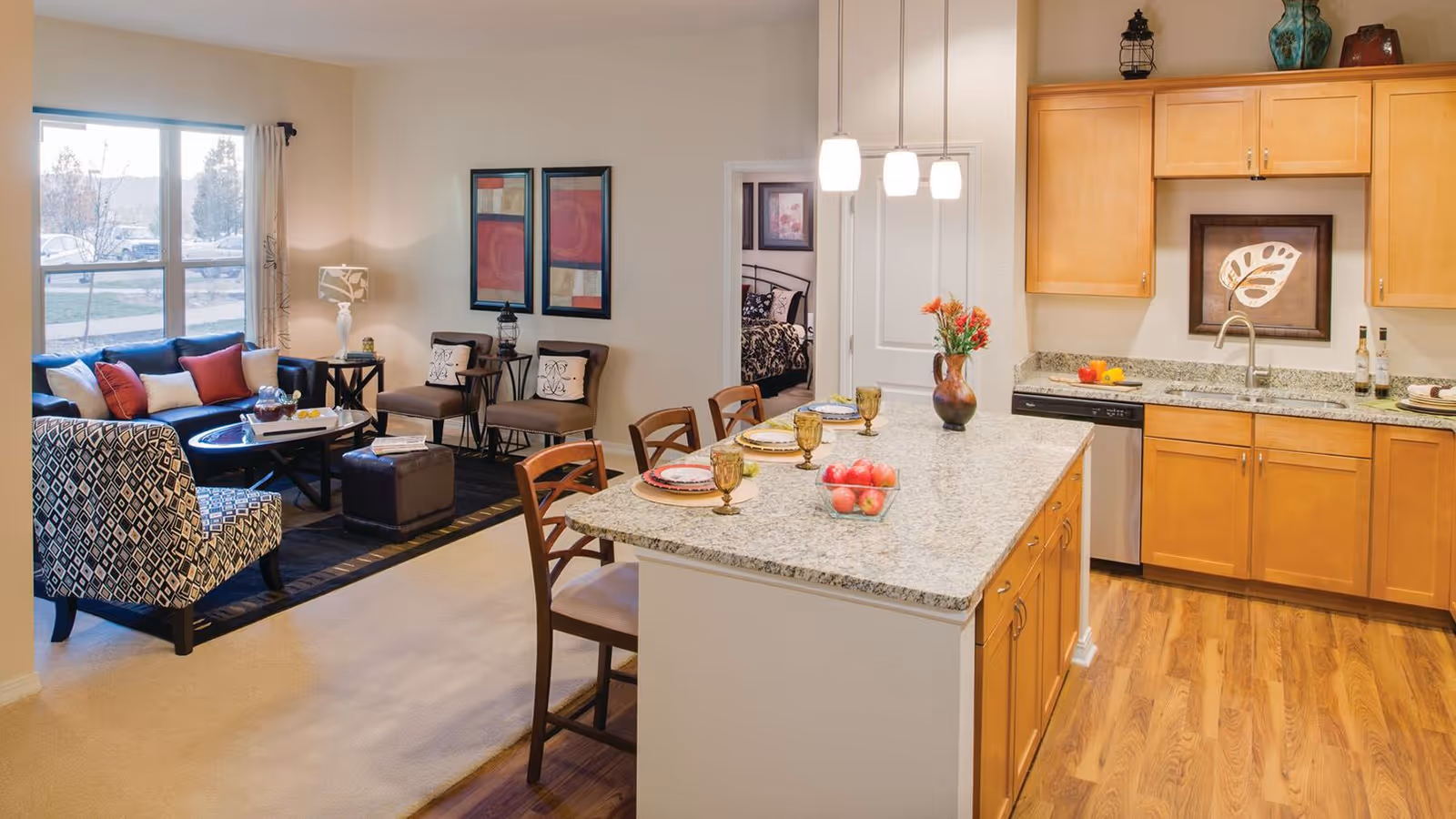 Interior view of a senior living community apartment showing a kitchen with wooden cabinets, granite countertops, and a kitchen island set with plates and glasses. Adjacent to the kitchen is a living room area with a patterned armchair, a dark blue sofa with red and white cushions, two beige chairs, a coffee table, and framed artwork on the wall. A bedroom is visible through an open doorway in the background.