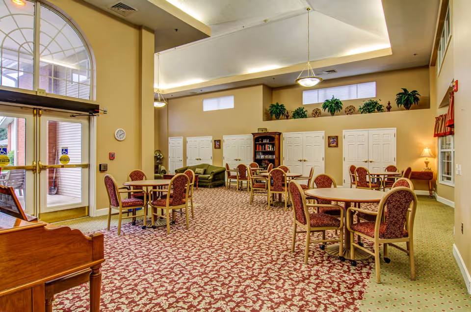 A spacious common area in a senior living facility with several round tables and upholstered chairs arranged on a patterned carpet. The room features high ceilings with recessed lighting, large windows near the ceiling, and a glass double door entrance. There is a green sofa, a wooden bookshelf with books, decorative plants on a ledge, and a small side table with a lamp. The walls are painted beige, and the room has a warm, inviting atmosphere.