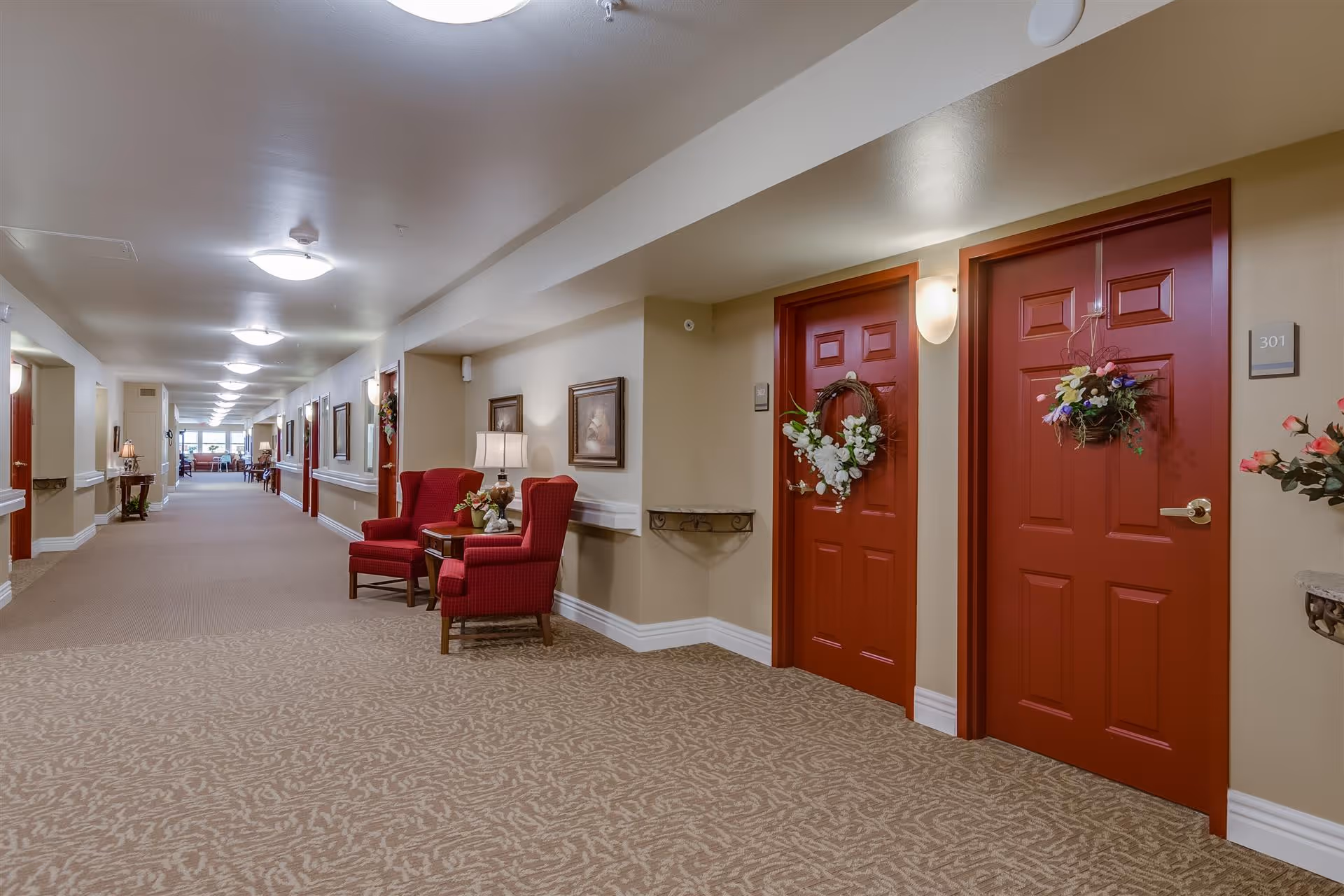 Carpeted interior hallway of an assisted living facility with red apartment doors decorated with wreaths and seating areas.