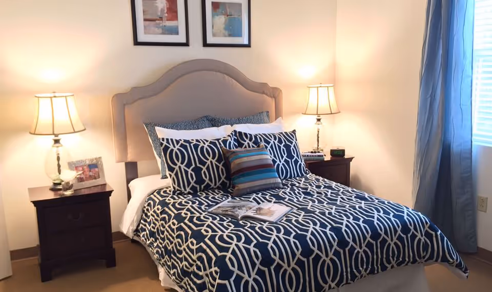 A neatly made bed with a beige upholstered headboard, decorated with multiple pillows in blue and white patterns and a small striped pillow in front. There are two dark wooden nightstands on either side of the bed, each with a lit table lamp. A book is open on the bed. The room has light-colored walls, two framed abstract paintings above the headboard, and a window with blue curtains on the right side.