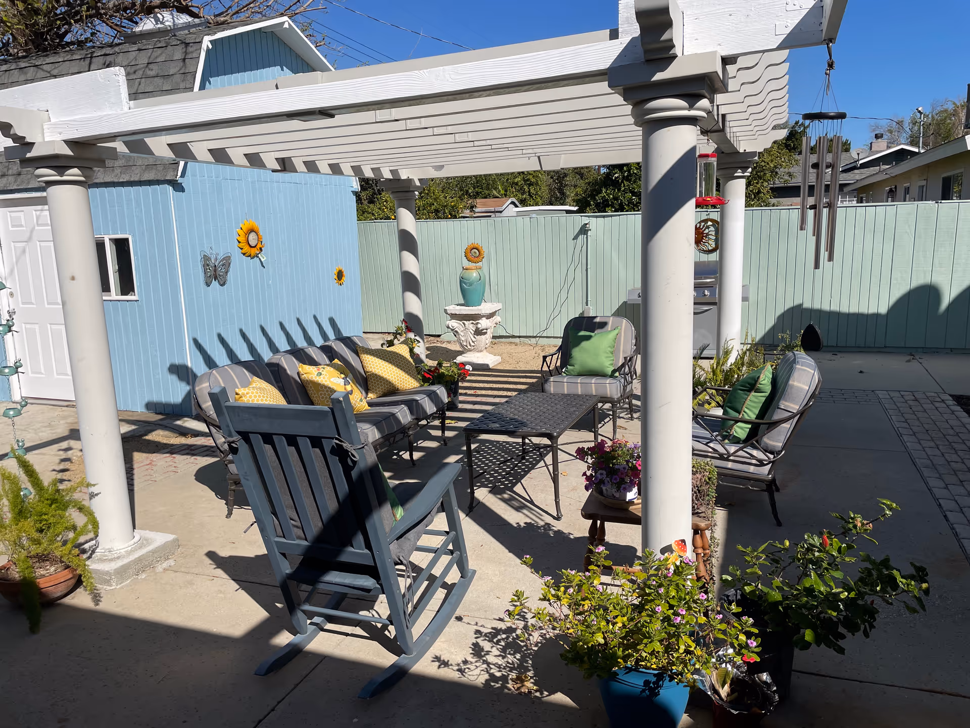 Outdoor patio area with a white pergola providing partial shade over a seating arrangement that includes a rocking chair, cushioned chairs, and a coffee table. The area is decorated with potted plants and flowers, and there is a blue shed with sunflower and butterfly wall decorations in the background. A wind chime hangs from the pergola.