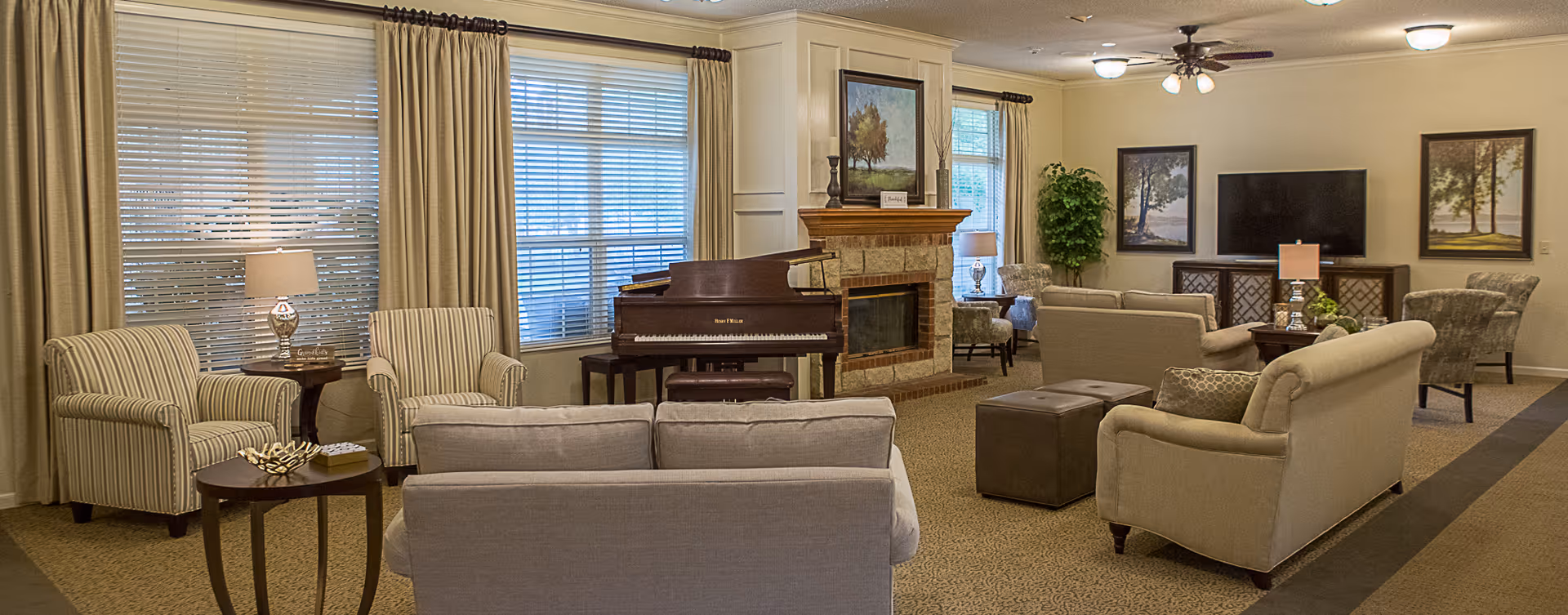 A spacious and well-lit living room with multiple beige and striped armchairs and sofas arranged around a central area. There is a grand piano near a stone fireplace with a painting above it. The room has large windows with blinds and curtains, framed artwork on the walls, a ceiling fan with lights, and various lamps providing warm lighting.
