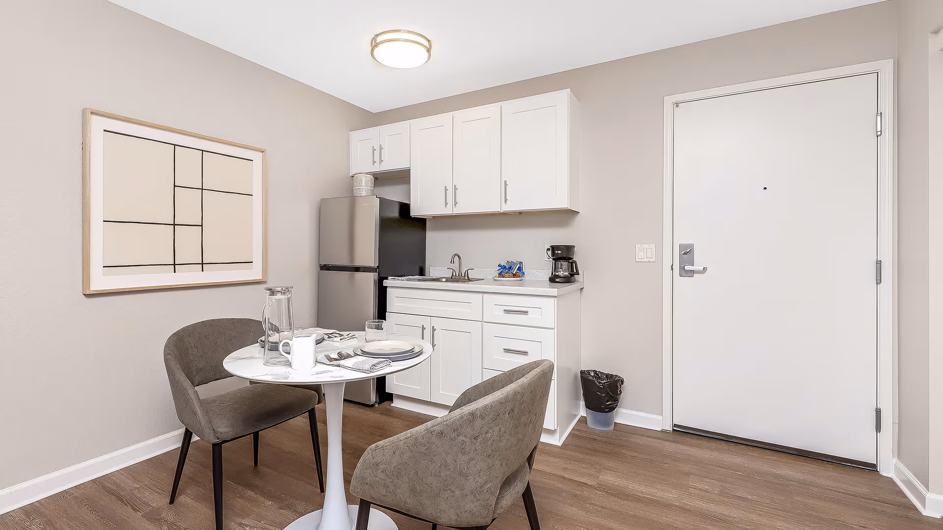 Small kitchen area with white cabinets, a stainless steel refrigerator, a sink, a coffee maker, and a small round dining table set for two with gray upholstered chairs. A modern abstract painting hangs on the wall, and a white door is visible to the right.