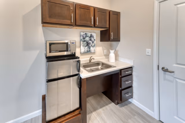 Small kitchenette area with a stainless steel mini refrigerator, a microwave on top, a sink with a faucet, dark wooden cabinets above and below the counter, a white pitcher on the counter, and a floral wall art hanging above the sink.