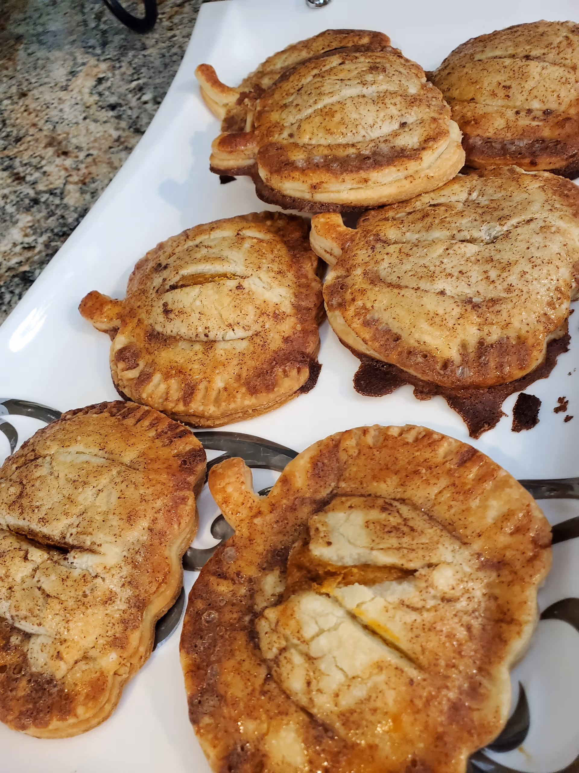A white platter holding several baked apple-shaped hand pies dusted with cinnamon on a granite countertop.