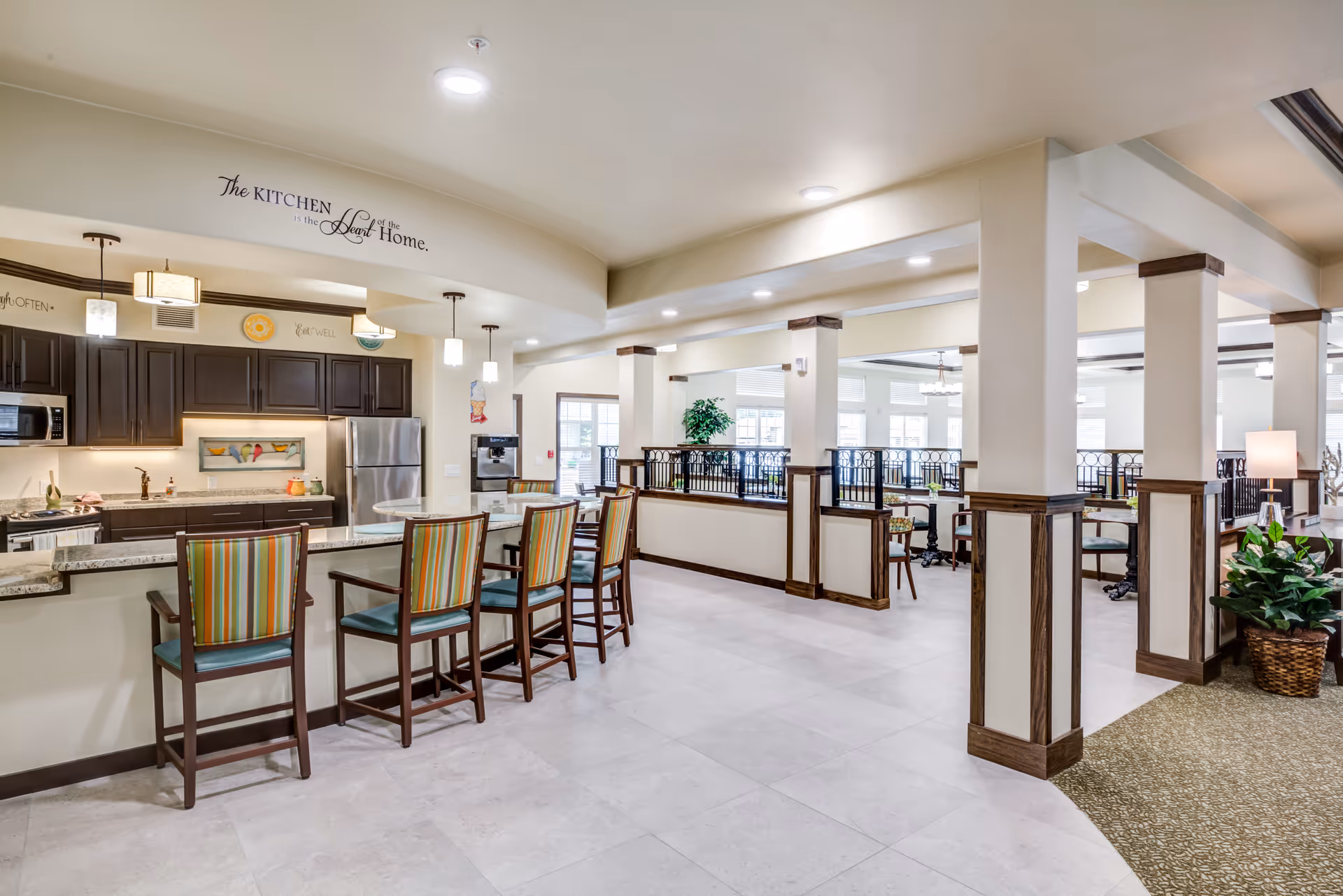 A spacious, well-lit kitchen area with a granite countertop bar and four high chairs with striped upholstery. The kitchen features dark wood cabinets, stainless steel appliances including a refrigerator and microwave, and pendant lights hanging from the ceiling. Adjacent to the kitchen is a dining area with tables and chairs, separated by half walls with decorative railings and columns. The walls are light-colored, and there are plants and lamps adding to the cozy atmosphere. A wall decal above the kitchen reads, 'The kitchen is the heart of the home.'