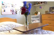 A bedroom in a senior living facility with a bed featuring white bedding, a wooden headboard, and a wooden overbed table holding a vase with colorful flowers. In the background, there is a wooden dresser, a plastic drawer unit, and a bulletin board with photos and notes on the wall.