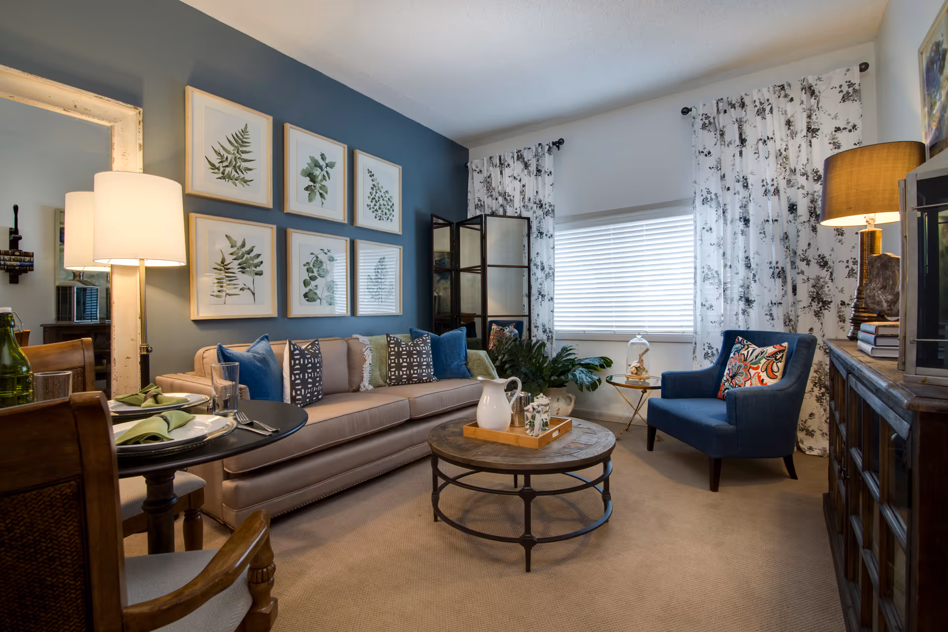 A cozy living room with a beige sofa adorned with multiple decorative pillows, a round wooden coffee table with a tray holding a white pitcher and cups, a blue armchair with a colorful pillow, and a wooden cabinet with a lamp on top. The walls feature framed botanical prints and a large mirror, with floral curtains covering a window.