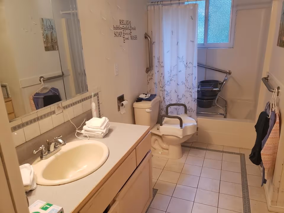 A bathroom with a sink and countertop on the left, a toilet with safety rails in the center, and a bathtub with a shower curtain and grab bars in the back. There are towels on the countertop and hanging on the wall. The floor is tiled, and there is a window above the bathtub letting in natural light. A wall decal with bathroom-related words is visible above the toilet.