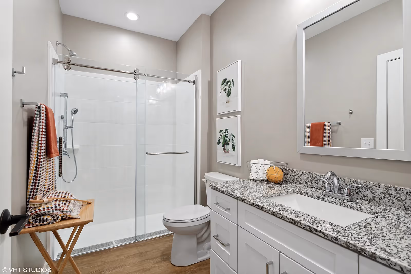 A modern bathroom featuring a glass sliding door shower with a handheld showerhead, a white toilet, and a white vanity with a granite countertop and a built-in sink. Above the vanity is a large rectangular mirror. Two framed botanical prints hang on the wall above the toilet. A wooden folding stool holds folded towels and a brush, and a towel hangs on a rack next to the shower. The floor has a wood-like finish and the walls are painted light beige.