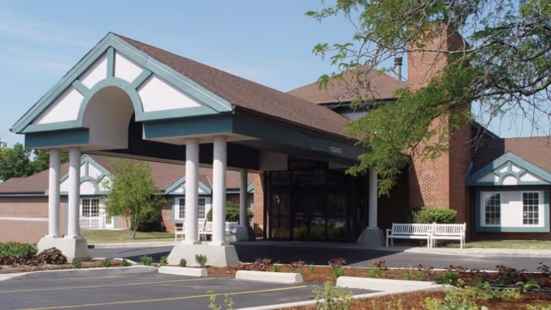 Front entrance of a brick senior living facility with a covered porte-cochère supported by columns, benches, and landscaped parking area.