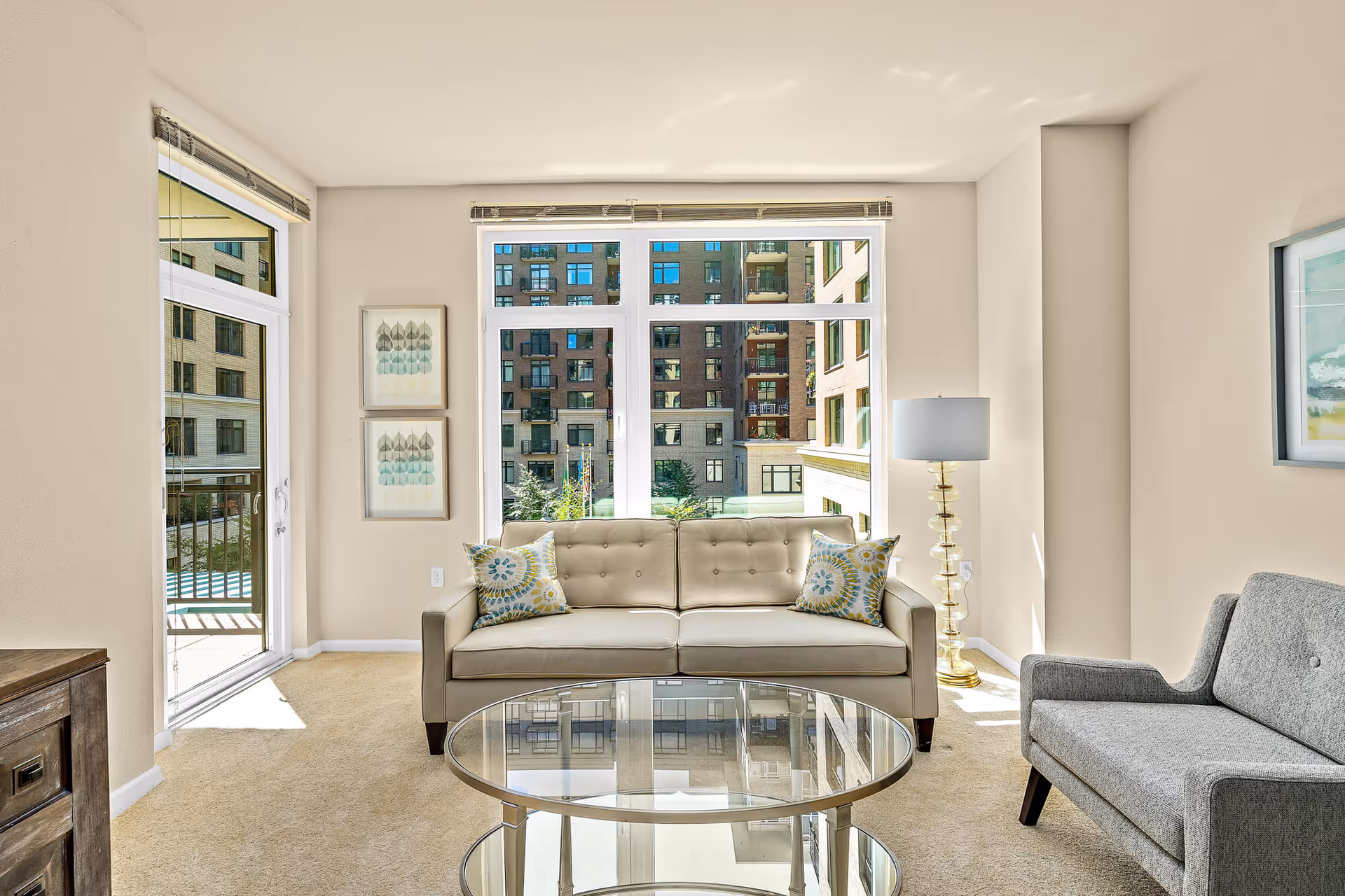 Bright living room with a beige tufted sofa and patterned pillows, glass coffee table, gray armchair, floor lamp, and large windows with a balcony door showing neighboring buildings.