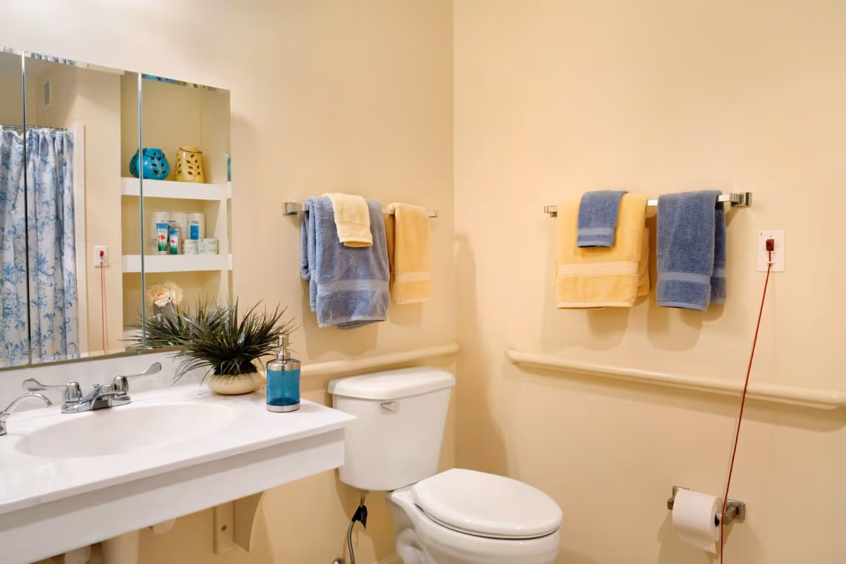 A clean bathroom with a white sink and countertop, a mirror above the sink, a toilet, and a shower with a blue and white patterned curtain. There are blue and yellow towels hanging on two towel racks on the walls. A small plant and a blue soap dispenser are on the sink countertop. The walls are painted a light beige color.