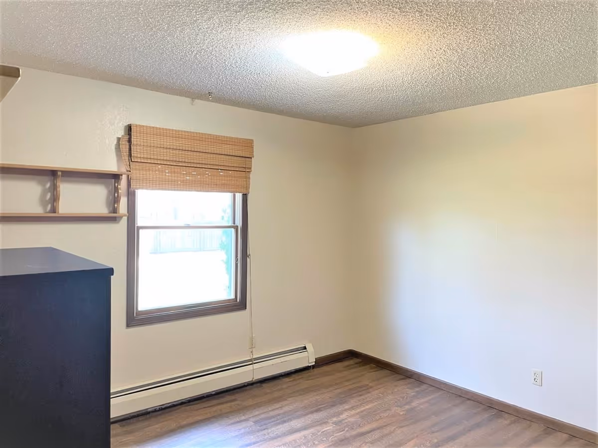 Empty room with wooden flooring, a window with bamboo blinds, a small wooden shelf on the wall, and a black dresser partially visible on the left side. The walls are painted light beige and there is a ceiling light fixture.