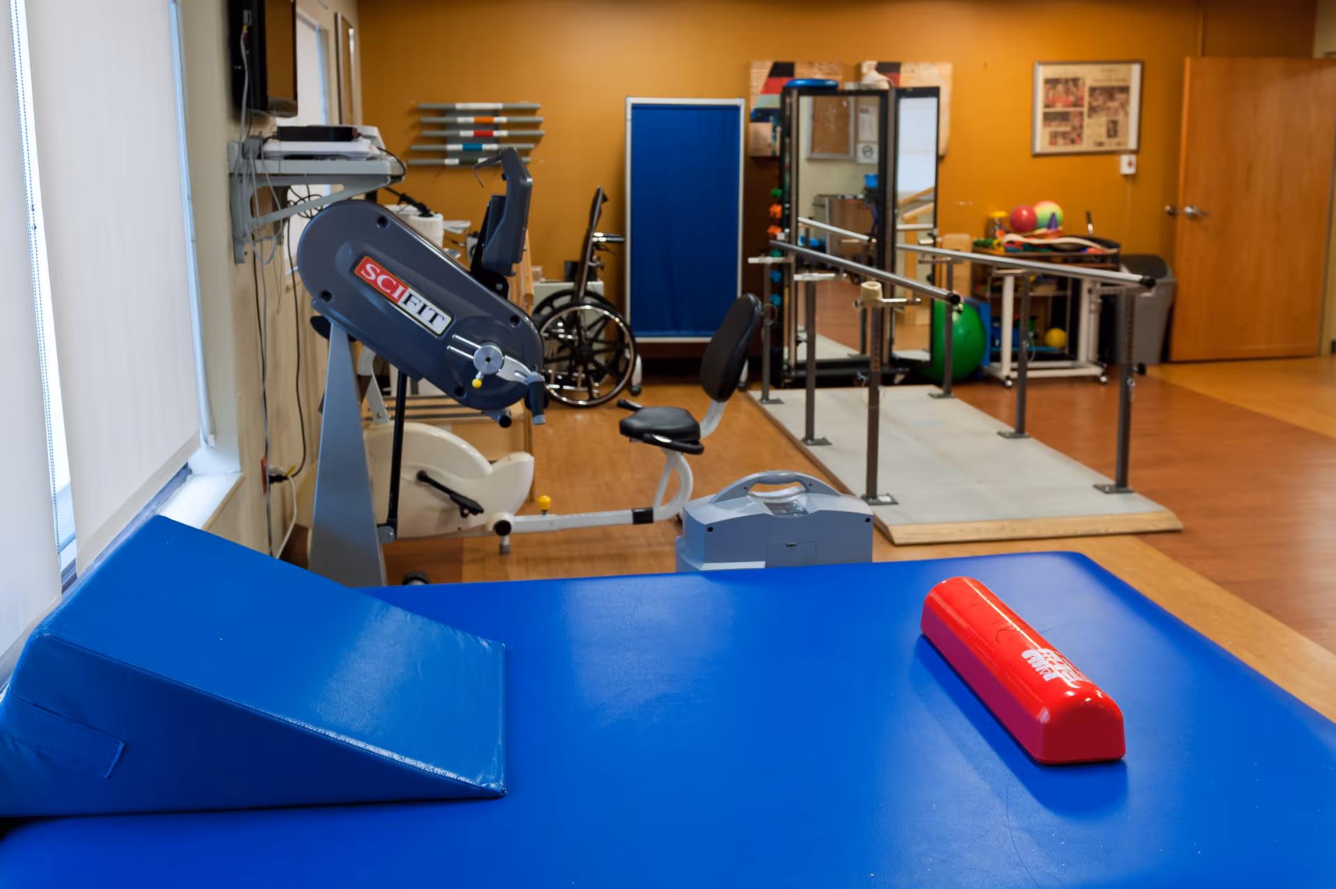 Physical therapy room with exercise equipment including a recumbent bike, parallel bars for walking practice, a wheelchair, a blue padded therapy table with a wedge cushion and a red cylindrical bolster, and various therapy balls and equipment in the background.
