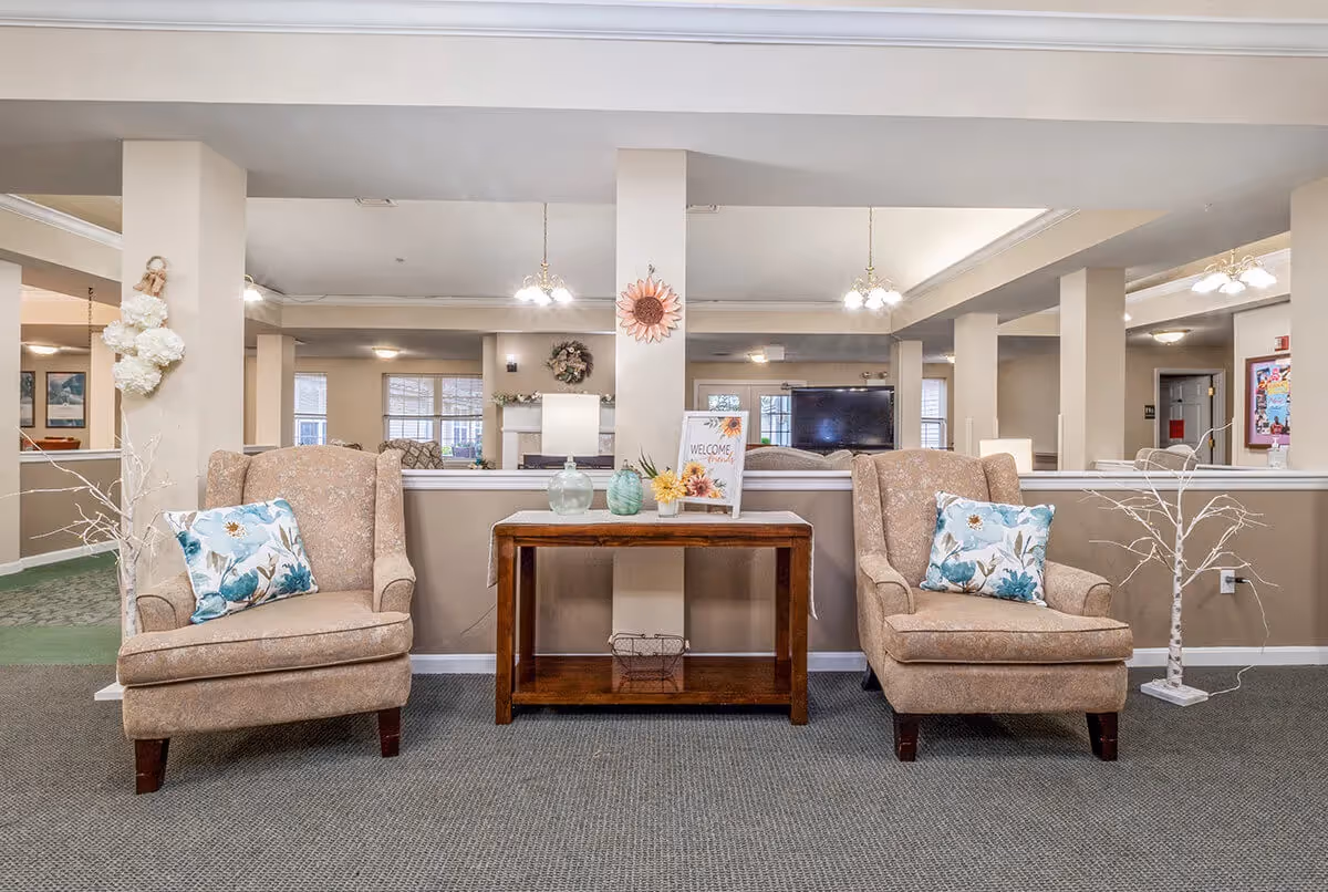 Two upholstered armchairs with floral pillows flank a wooden console table in a spacious senior living room lounge.