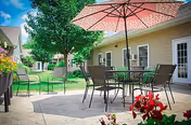 Outdoor patio area at a senior living facility with metal tables and chairs under a large pink umbrella, surrounded by green grass, trees, and beige building walls with windows and doors.