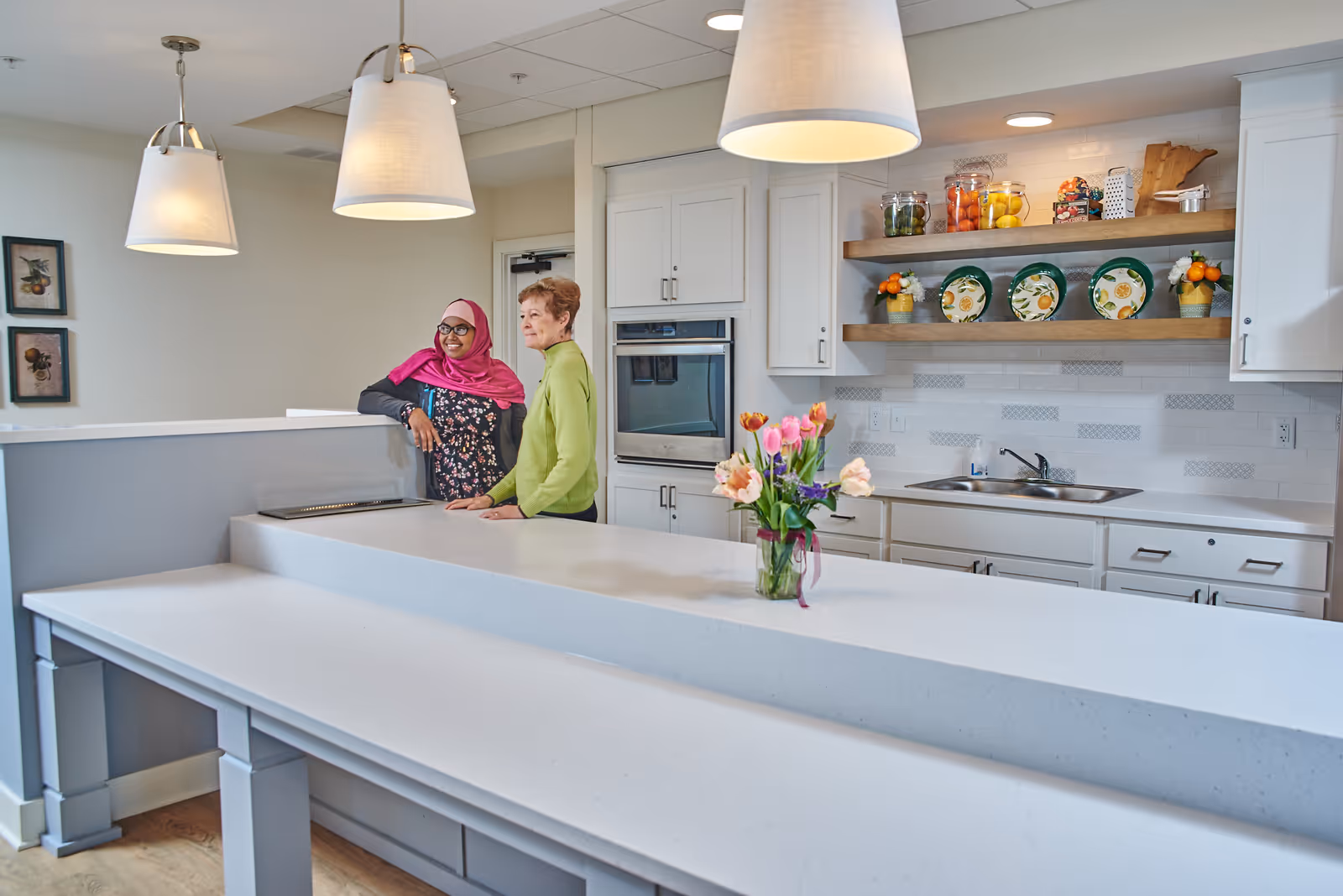 Two women standing and smiling in a modern kitchen area with white countertops, white cabinets, a built-in oven, a sink, and decorative shelves holding plates and jars. There are three pendant lights hanging from the ceiling and a vase with colorful flowers on the counter.
