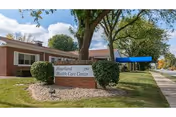 Brick senior living facility exterior with sign, trees, and a sidewalk.