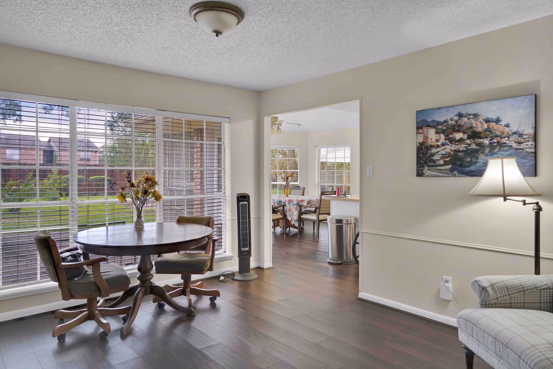 A bright interior room with large windows covered by white blinds, featuring a round wooden table with two cushioned chairs on casters. A vase with flowers sits on the table. The room has dark wood flooring and beige walls. In the background, there is an adjoining dining area with a table covered by a floral tablecloth and several chairs. A floor lamp and a painting of a coastal town hang on the wall to the right.