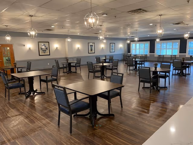 A spacious dining room with multiple square tables and chairs arranged neatly on a polished wooden floor. The room is well-lit with hanging pendant lights and wall sconces. There are framed artworks on the walls and large windows with white shutters letting in natural light.