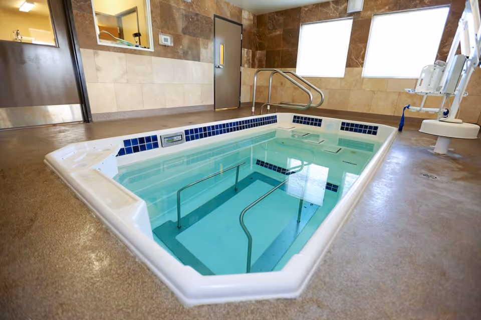 Indoor therapeutic pool with handrails and steps for easy access, surrounded by tiled walls and two large frosted windows allowing natural light. There is a pool lift chair on the right side for accessibility.