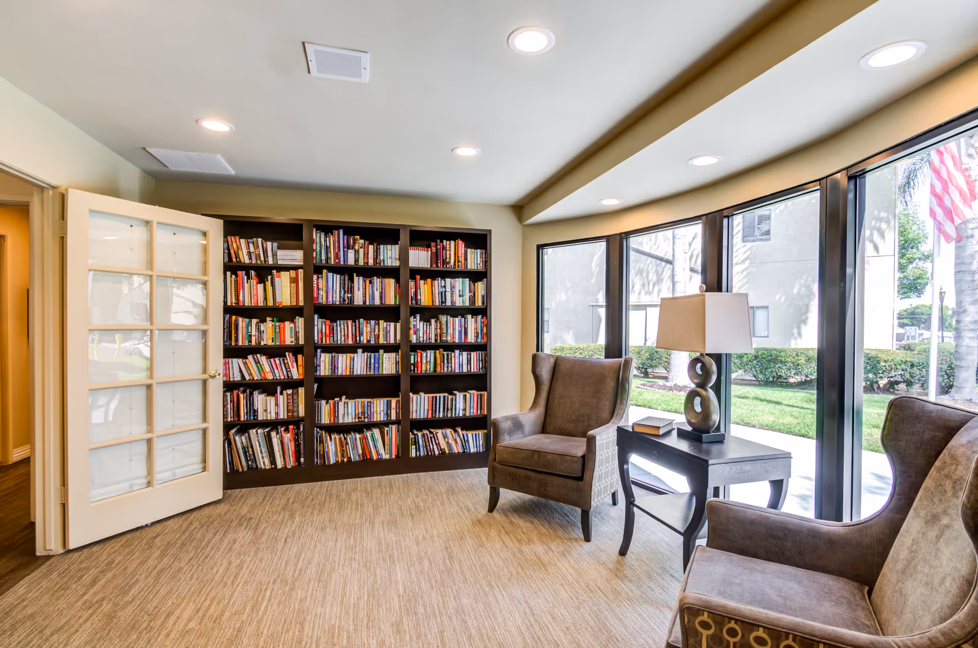 A cozy reading area in Canterbury Court Senior Apartments featuring a large bookshelf filled with books, two upholstered armchairs, a side table with a lamp and a book, and large windows letting in natural light with a view of greenery outside.