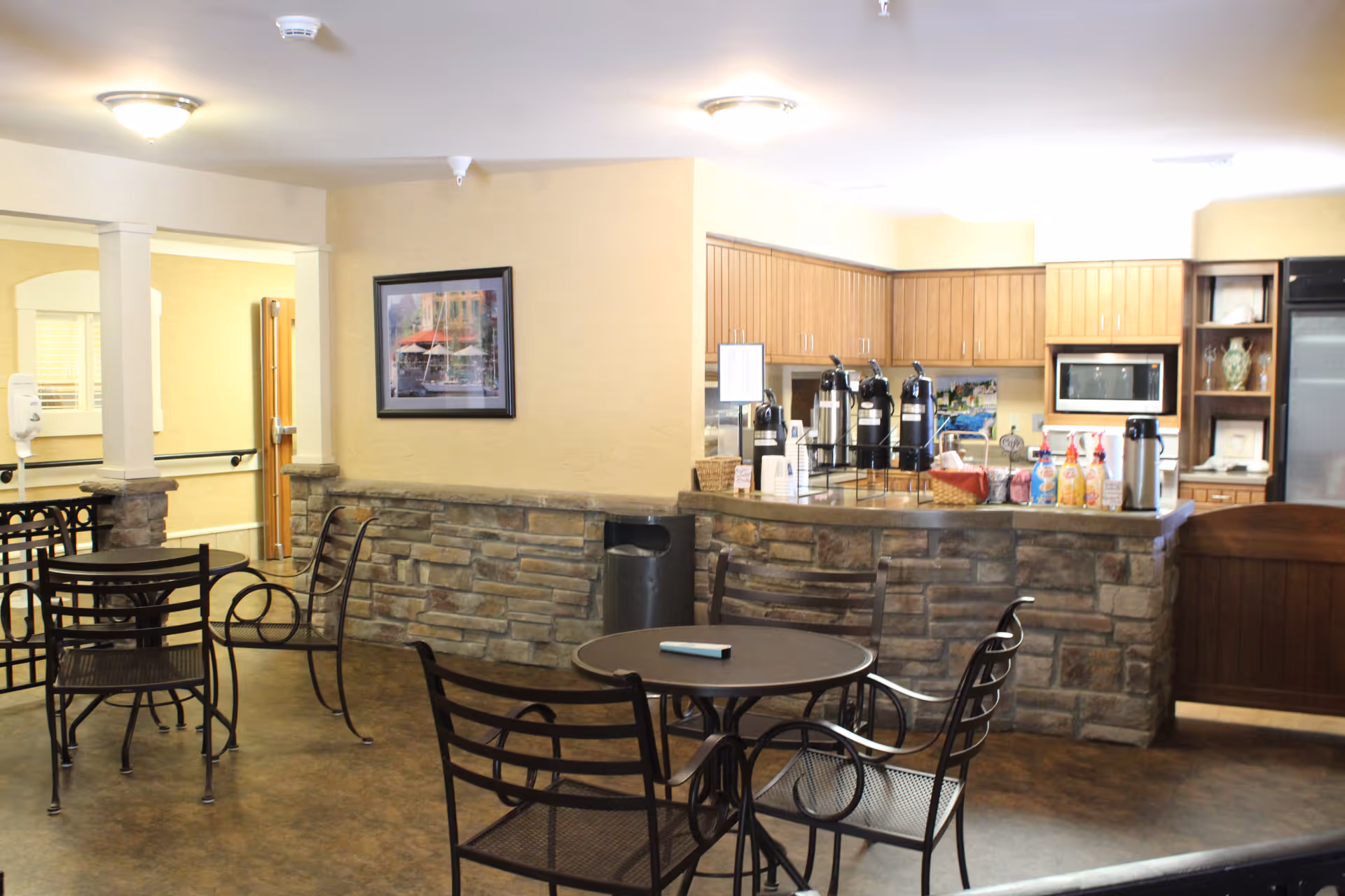 A cozy common area with a stone-faced counter and wooden cabinets in the background, featuring coffee dispensers and various condiments on the counter. There are several round metal tables with matching chairs arranged in the foreground. The walls are painted beige, and a framed picture hangs on one wall. The area is well-lit with ceiling lights.