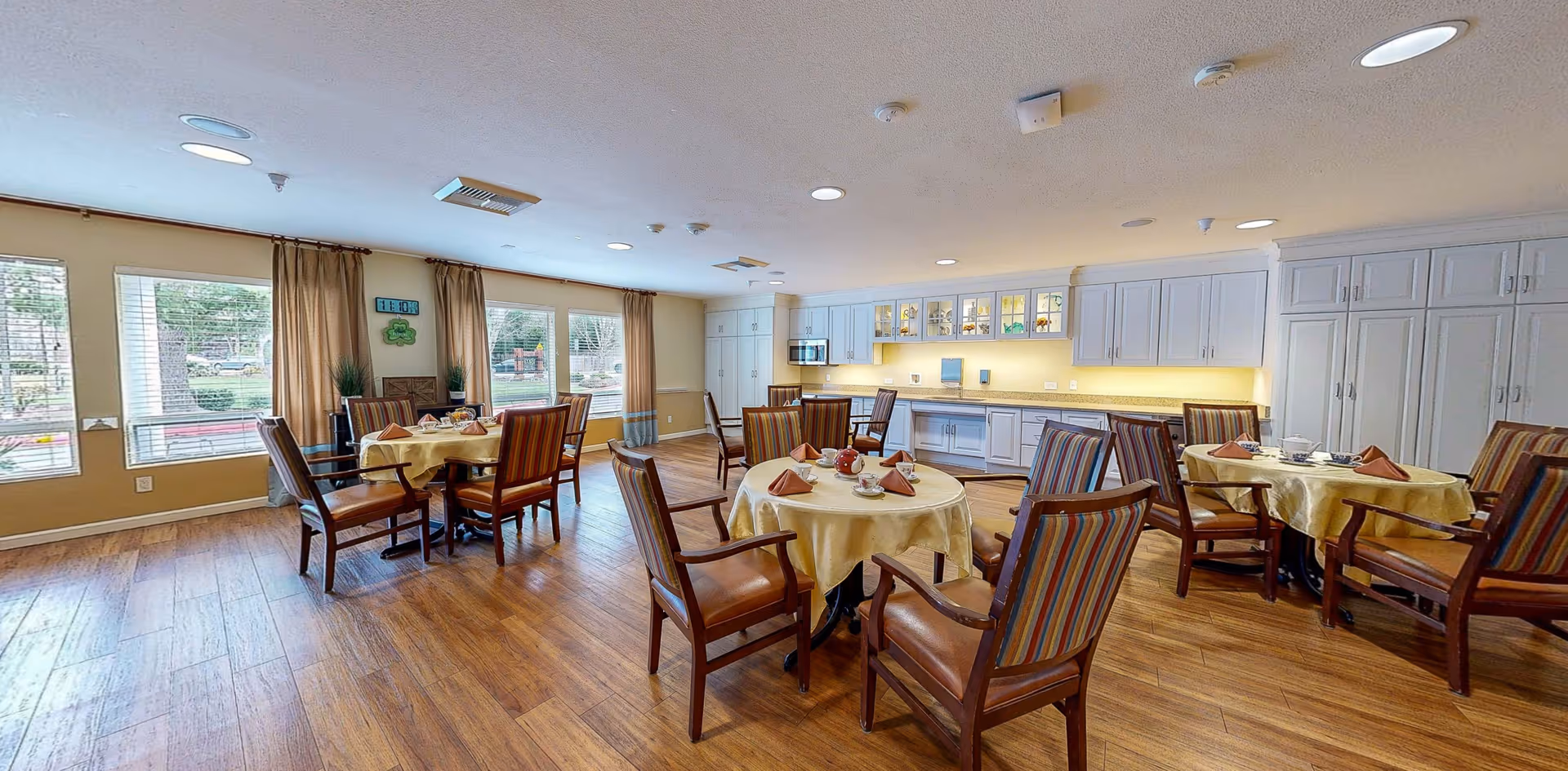 Bright memory-care dining room with round tables covered in yellow tablecloths, wooden chairs, large windows, and a long cabinet/kitchenette wall.