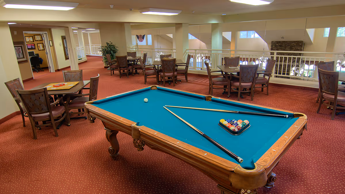 A spacious senior living common area with a pool table in the center surrounded by tables and chairs on red carpet.