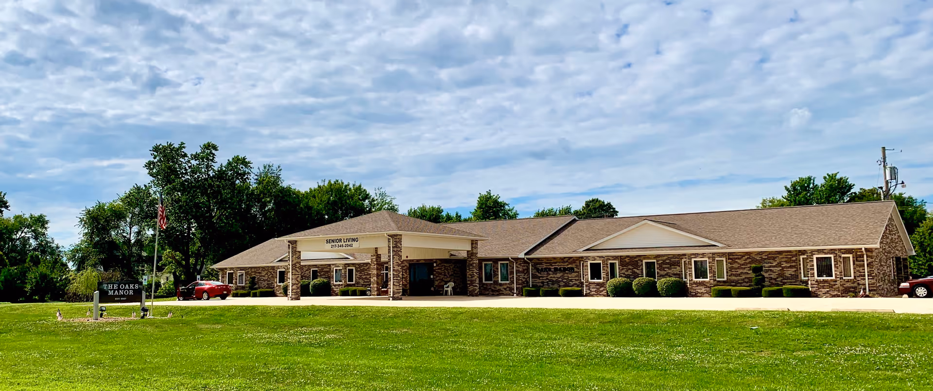 Exterior view of The Oaks Manor senior living facility, a single-story brick building with a covered entrance and a well-maintained lawn in front. There is a sign on the lawn that reads 'The Oaks Manor' and an American flag on a flagpole nearby. The sky is partly cloudy.