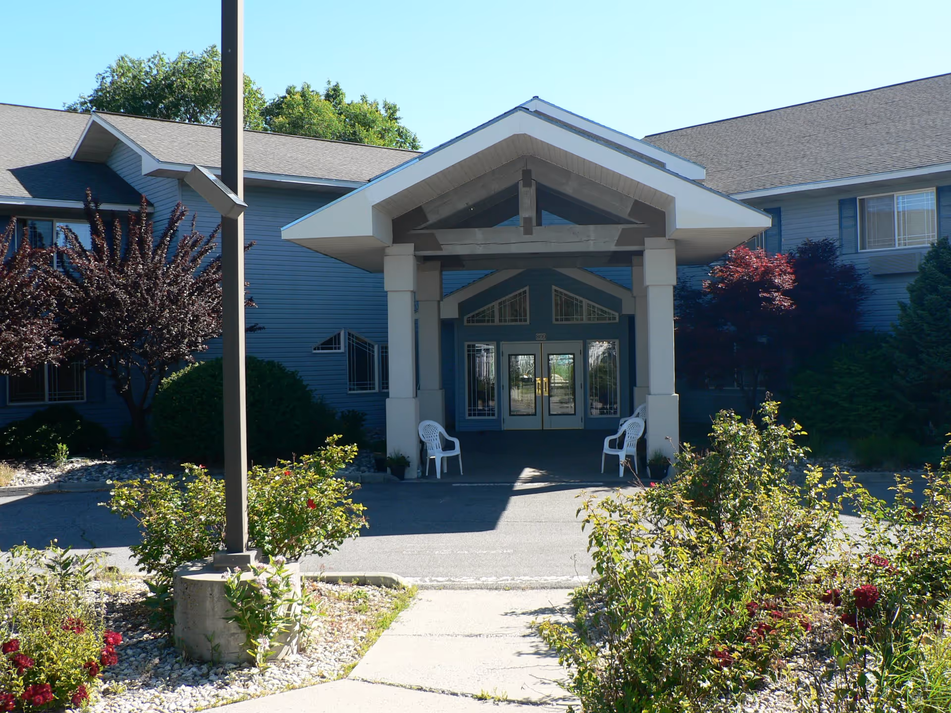Front entrance of a retirement and assisted living facility with a covered porch supported by columns, two white plastic chairs on either side of the entrance, surrounded by bushes and trees under a clear blue sky.
