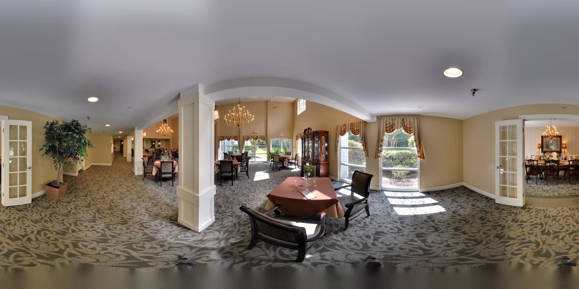 A spacious and elegant dining area in a senior living facility with multiple tables and chairs arranged neatly. The room features large windows with draped curtains allowing natural light to fill the space. There are chandeliers hanging from the ceiling, and a china cabinet is visible against one wall. The carpet has a decorative pattern, and there are French doors leading to an adjacent room with a similar dining setup.
