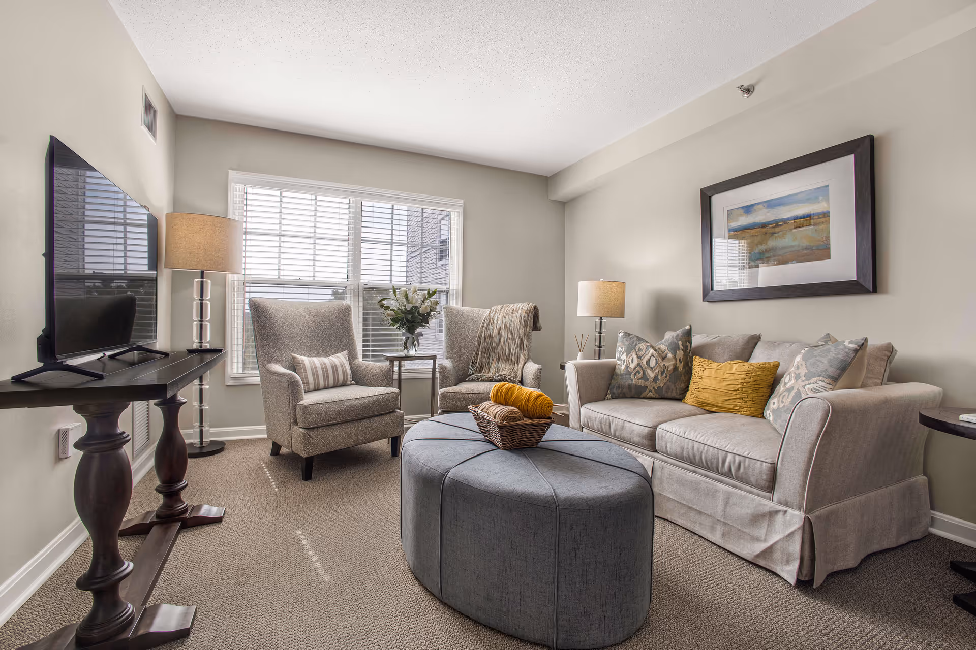 A cozy living room with a beige sofa adorned with patterned and solid yellow pillows, two upholstered armchairs with a small table holding a vase of flowers between them, a large gray ottoman in the center with a basket on top, a flat-screen TV on a dark wooden stand, two table lamps, and a framed landscape painting on the wall.